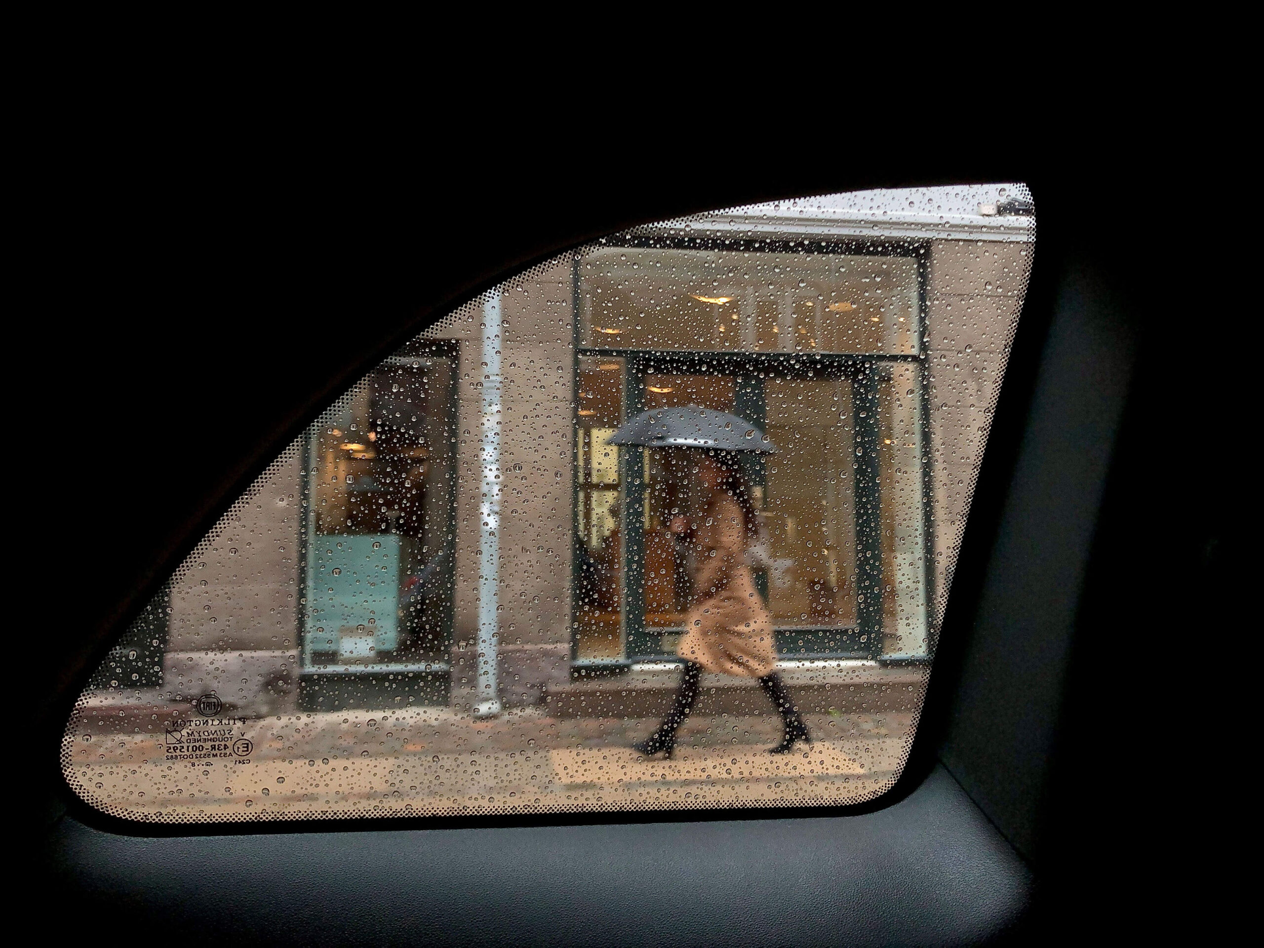 Woman in beige coat walking with an umbrella in the rain in Copenhagen seen through rain drops on the rear window of a car.