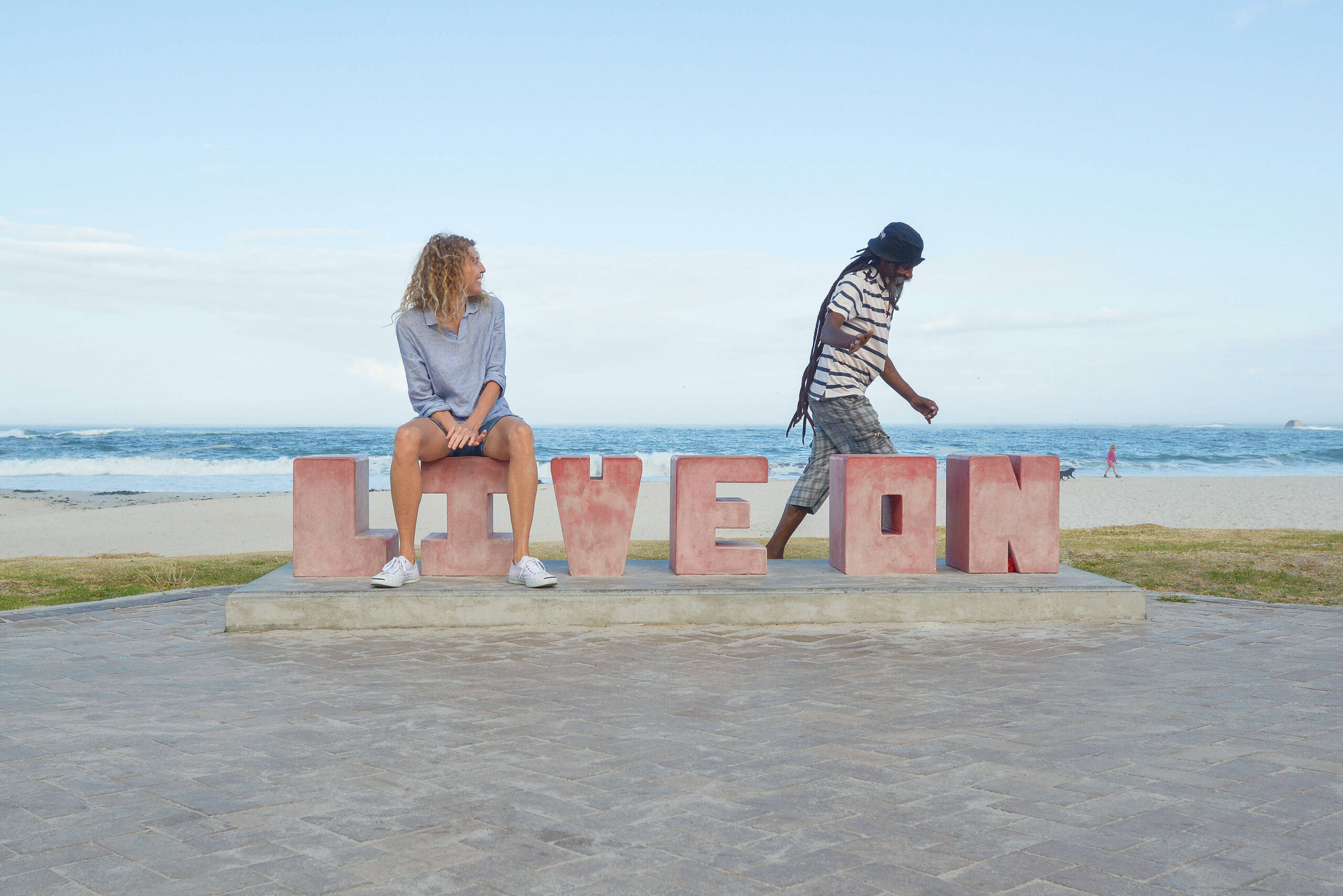 A Rastafarian man walks past a young woman seated on a sign that says "live on" in Camps Bay, South Africa. Their quick connection created one of those heart moments to be remembered.