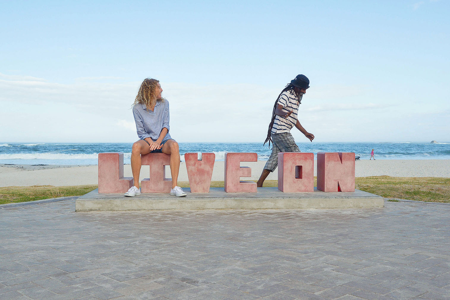 Camps Bay Beach sculpture, Live On, with happy woman seated and man greeting her in casual passing in Cape Town, South Africa.