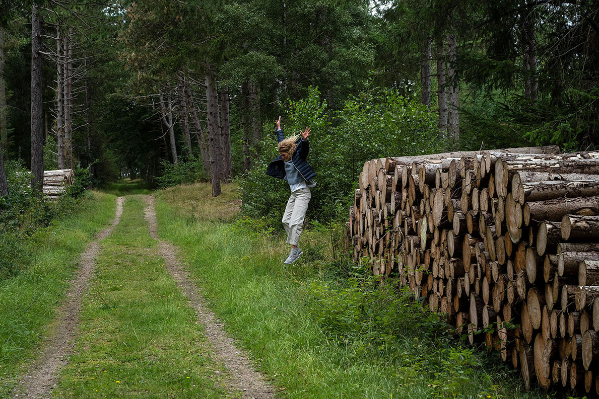 Woman jumping down from a stack of cut tree logs in a pine forest in Denmark.