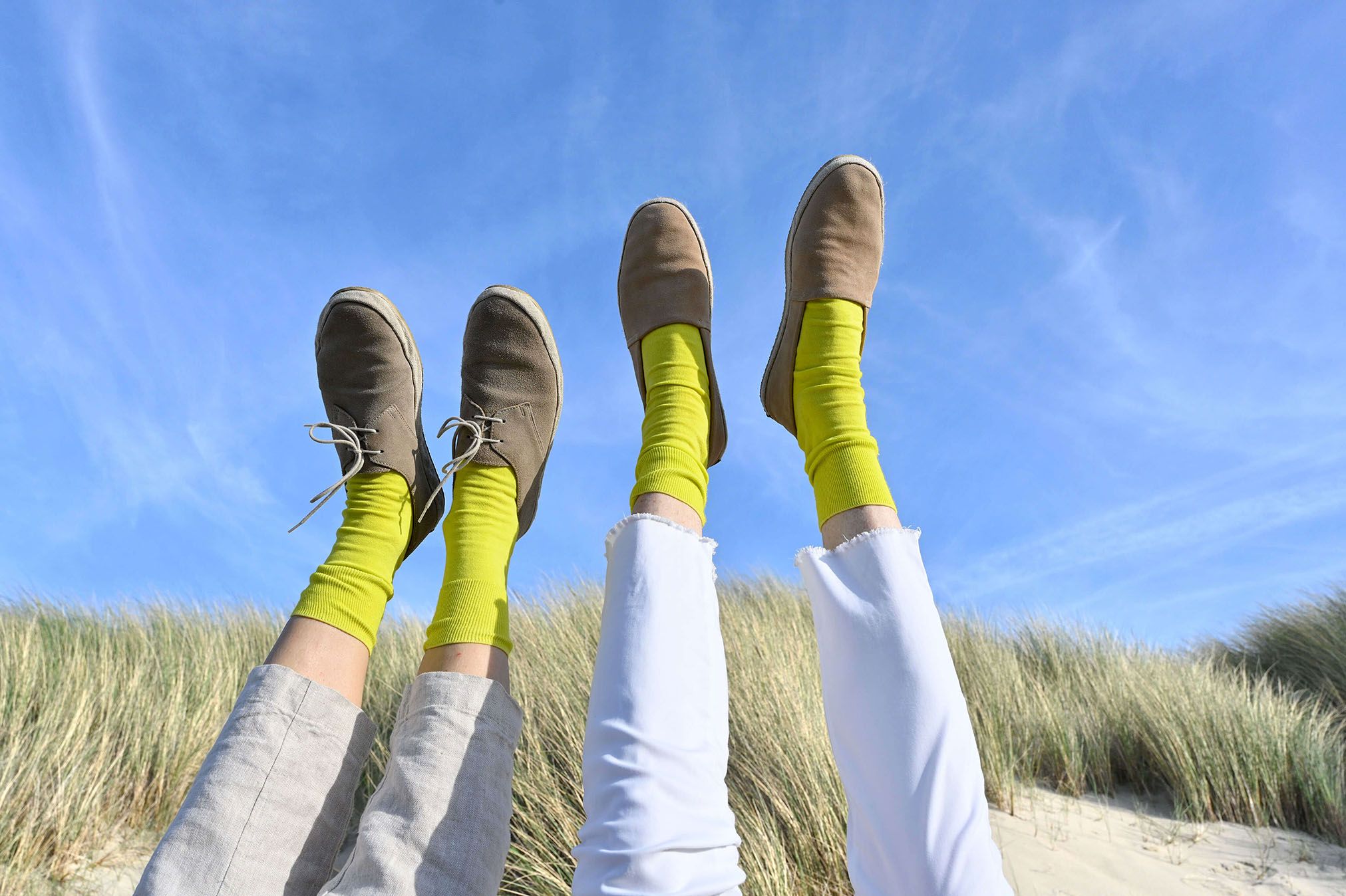 Two pairs of feet in yellow socks and brown suede shoes against a blue sky and dunes in Zeeland in the Netherlands